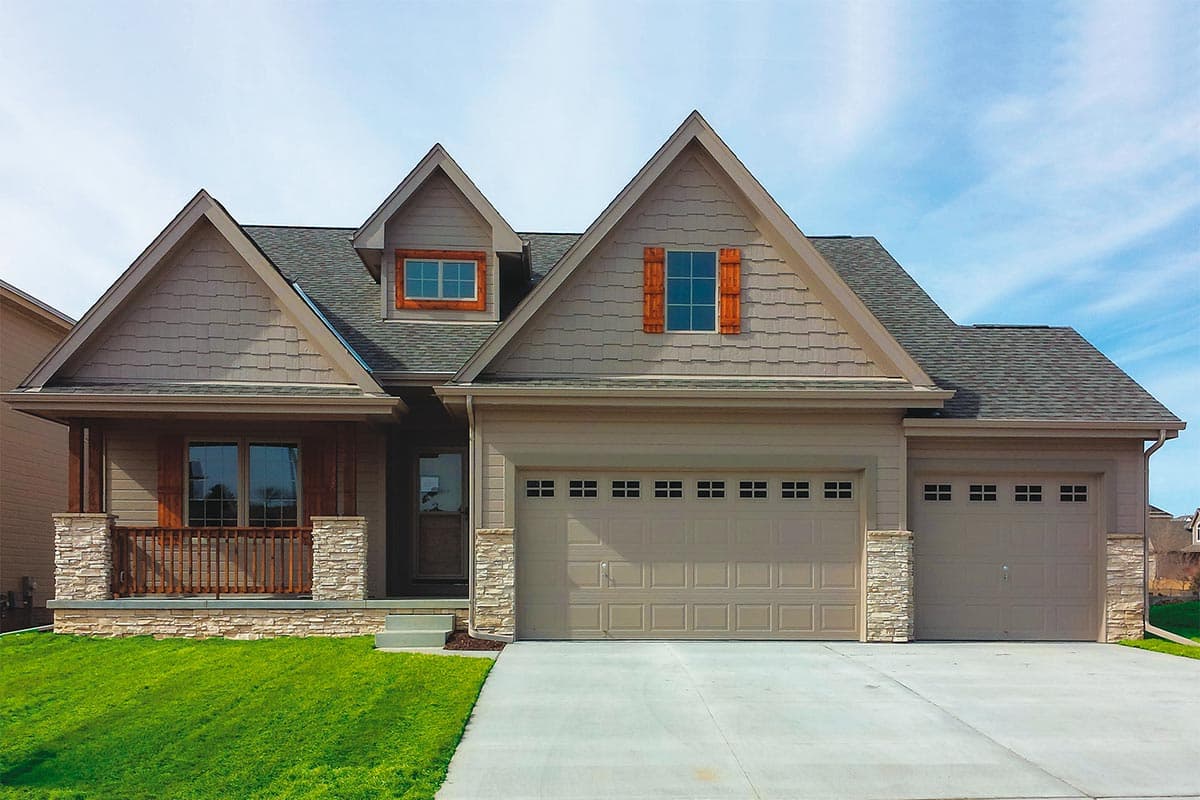 House plan exterior. Craftsman style, single-story home with a porch, gables, and a two-car garage. Features stone facade and dormer window.