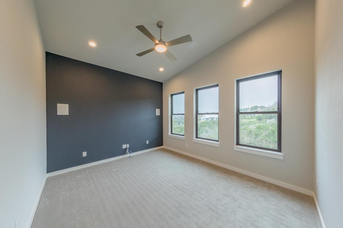 Interior view of a bedroom with a vaulted ceiling, ceiling fan, and three large windows overlooking a landscape.