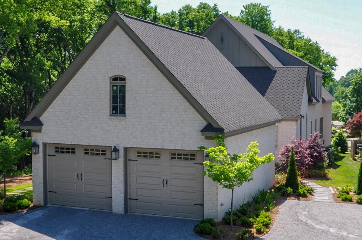 House plan exterior with light brick facade, a prominent gable, arched window, and a three-car garage with carriage-style doors.