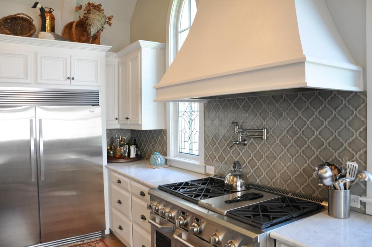Kitchen interior with stainless steel refrigerator, gas range, white cabinetry, pot filler, and decorative tile backsplash.