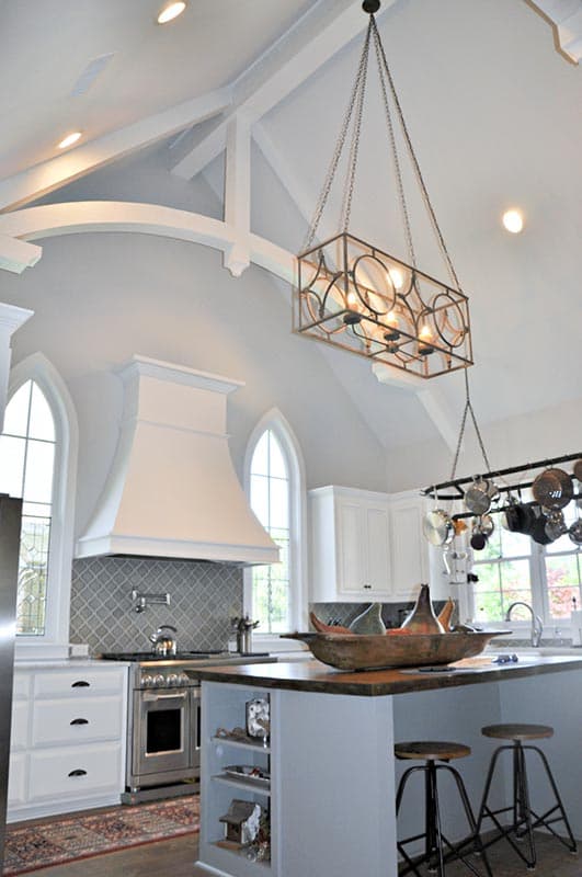 Kitchen with vaulted ceiling, exposed beams, custom range hood, arched windows, island with seating, and hanging pot rack.