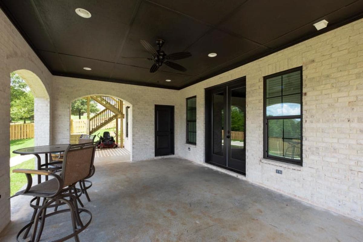 Covered outdoor living space with white brick walls, black ceiling fan, sliding glass doors, and access to an outdoor staircase.