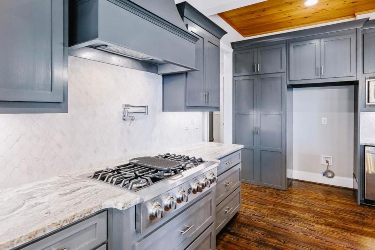 Kitchen with gray cabinets, white tile backsplash, gas range, marble countertops, and wood flooring.