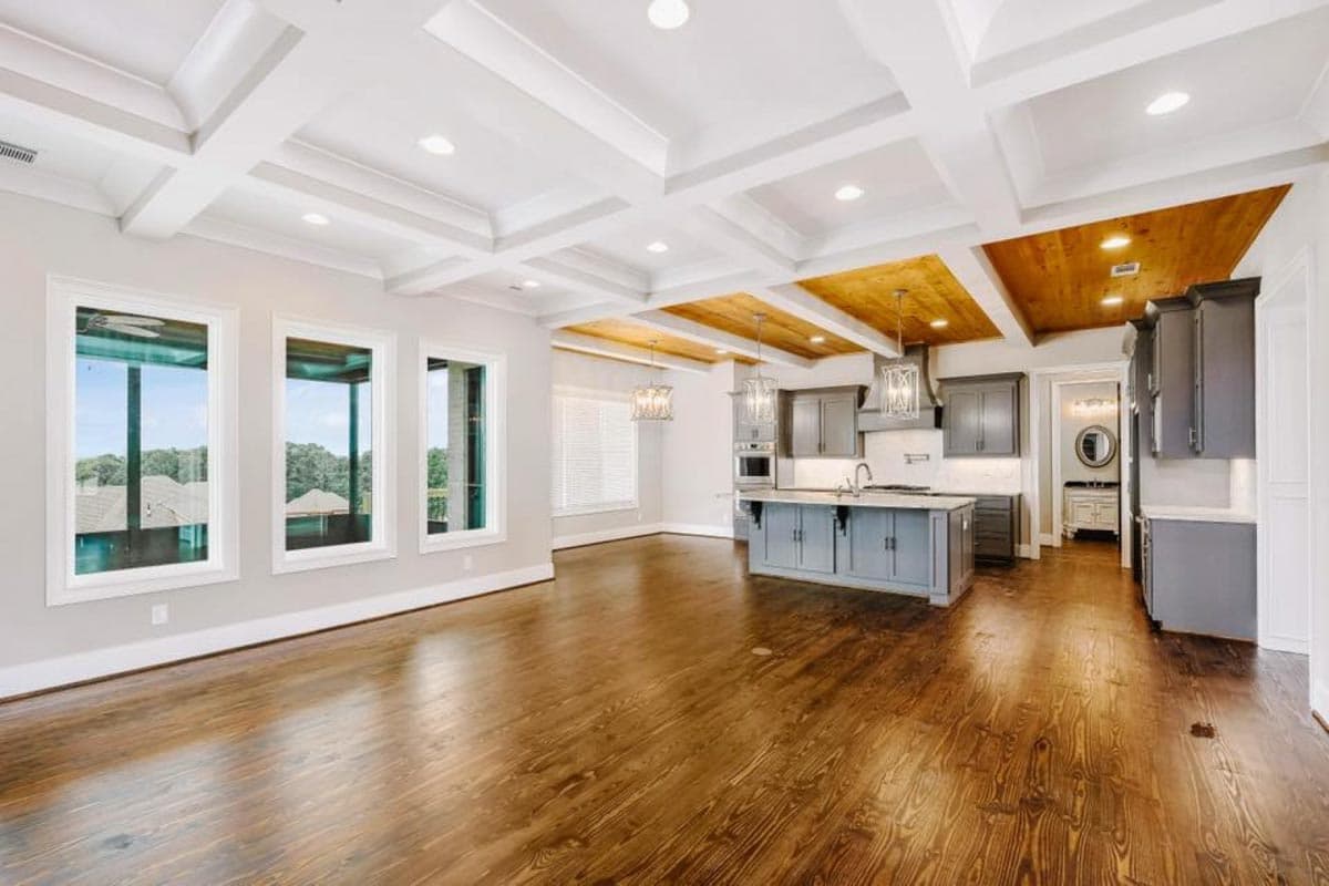 Open-concept living area with coffered ceiling, wood accents, kitchen island, and three large windows.