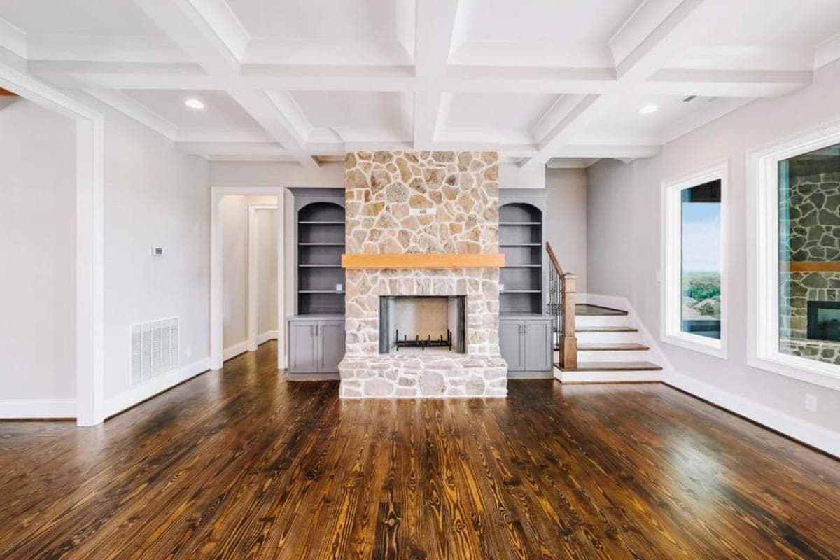 Interior view of living room with stone fireplace, built-in bookshelves, coffered ceiling, hardwood floors, and staircase.