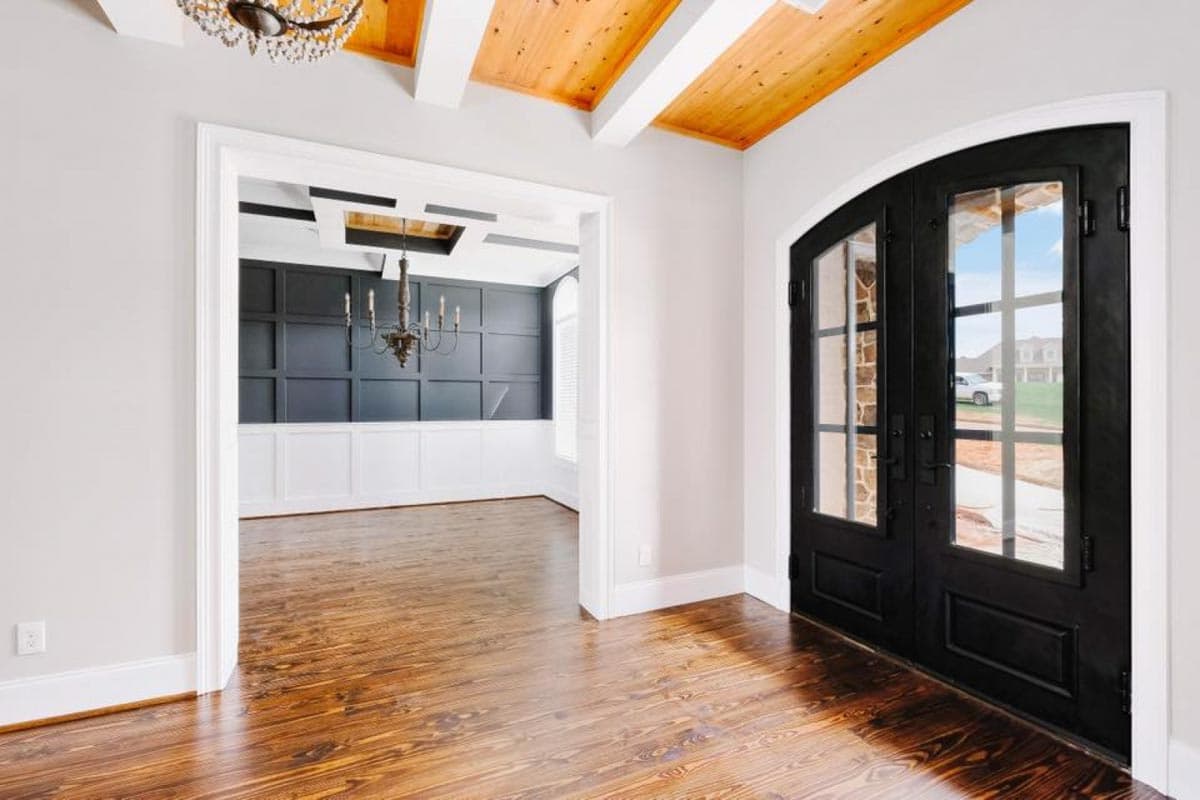 Interior view showing wood-paneled dining room with chandelier, visible through doorway from foyer with double doors and coffered ceiling.