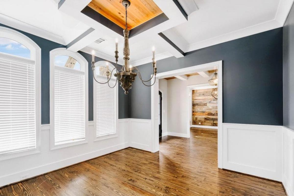 Dining room with coffered wood ceiling, arched windows with blinds, wainscoting, and chandelier. Opens to a wood-paneled room.