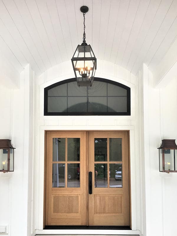 House plan exterior: Double wooden doors with glass panes, arched transom window, and two lantern sconces below a large hanging pendant.