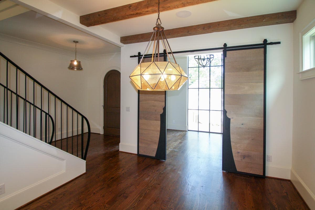 Interior view of a home with a grand staircase, decorative chandelier, wooden barn doors, and exposed ceiling beams.