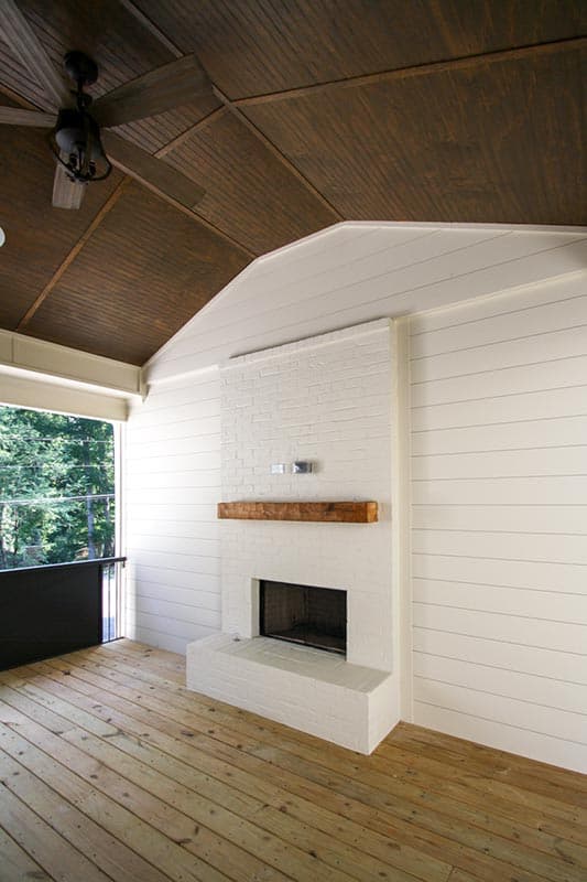 Covered porch with wood plank ceiling, fan, shiplap walls, white brick fireplace, and wood floors.
