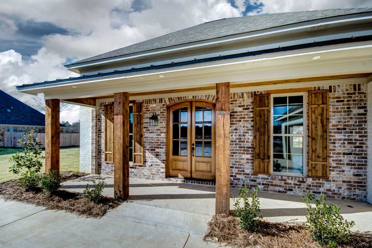 House plan exterior with brick facade, covered porch, wood beams, arched double entry doors, and wood shutters.