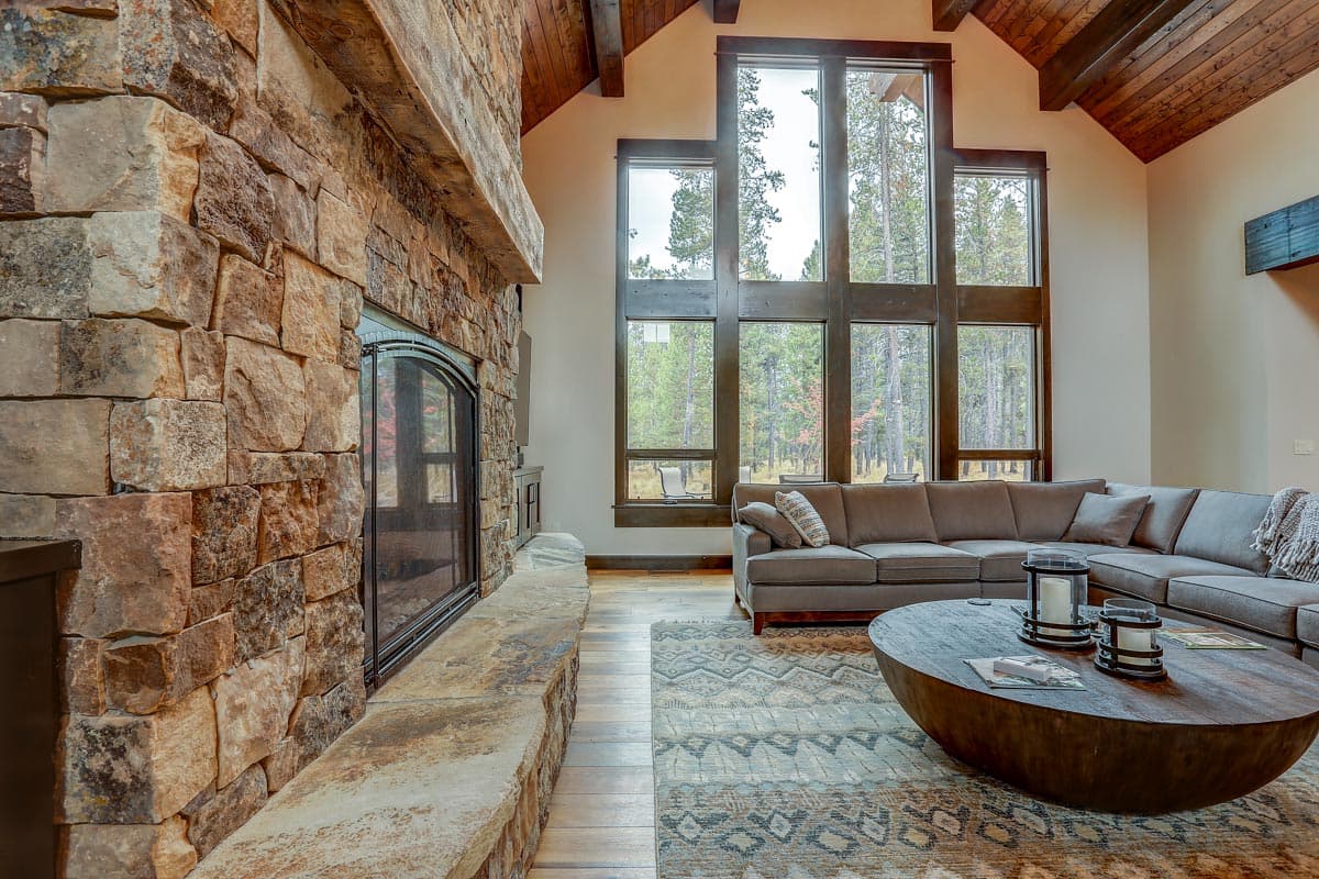 Great room interior with stone fireplace, vaulted wood beam ceiling, and large windows overlooking a forest.