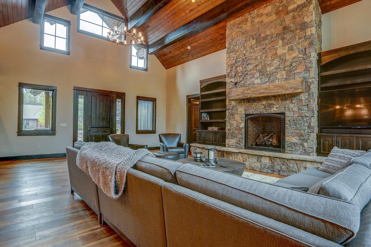 Interior view of a great room with vaulted wood beam ceiling, stone fireplace, built-in shelving, and large windows.