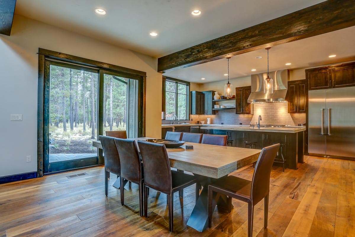 Dining area and kitchen with island, stainless steel appliances, dark wood cabinetry, and large sliding glass doors overlooking a forest.