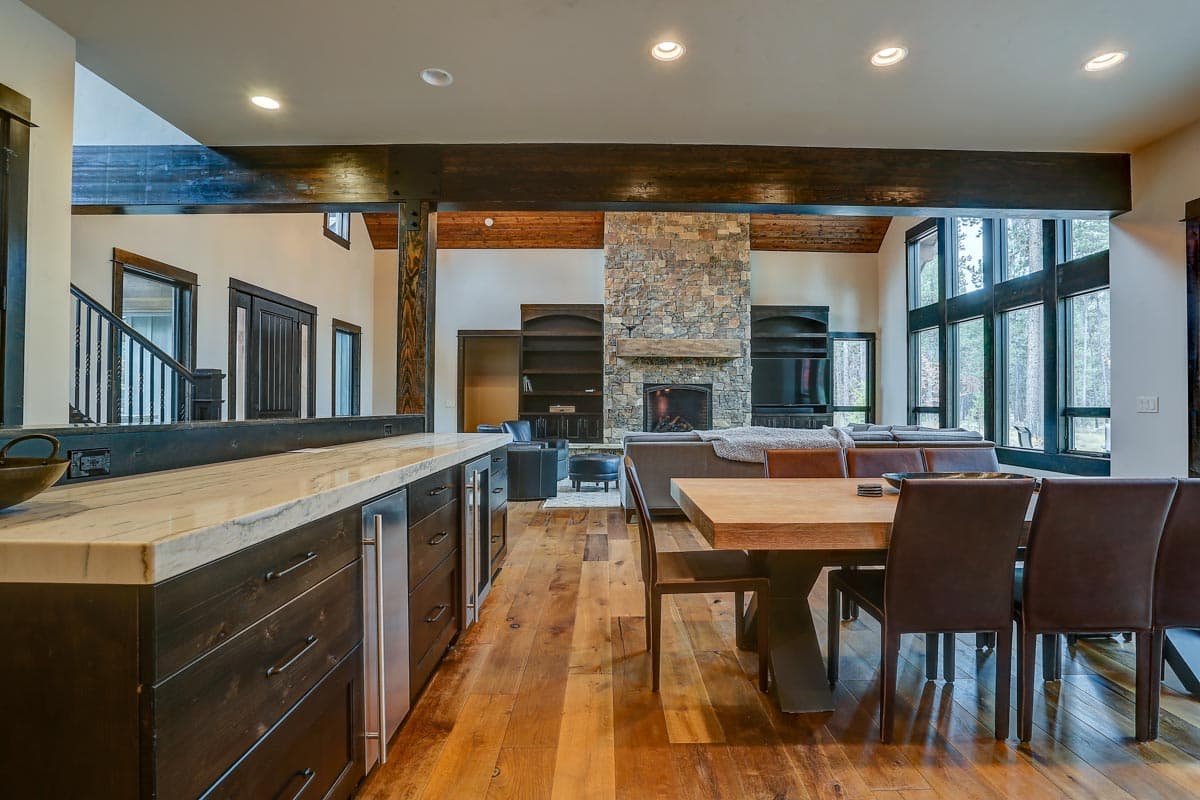Open concept living area with stone fireplace, large windows, dining table, and kitchen island with dark wood cabinetry.