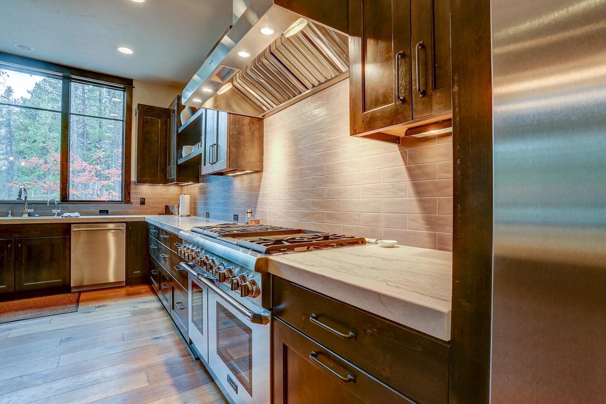 Kitchen featuring a stainless steel range with a prominent hood, marble countertops, dark wood cabinetry, and a large window overlooking trees.