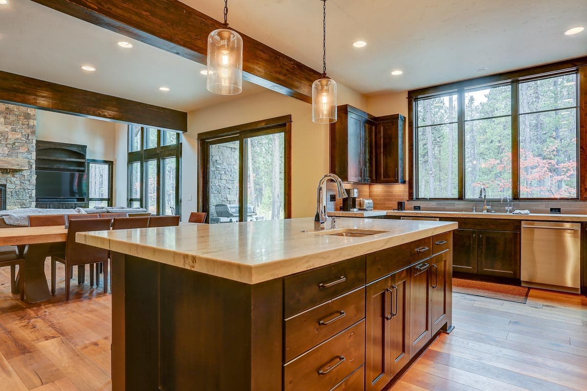Kitchen with large island, stone fireplace, wood beams, and forest view through expansive windows.