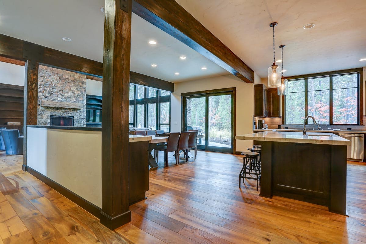 Interior view of an open-concept living area with a stone fireplace, dining table, kitchen island with pendant lights, and large windows.