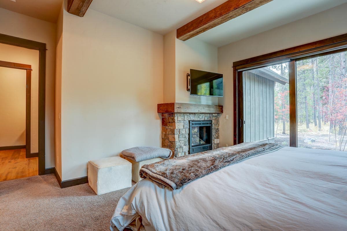 Primary bedroom with stone fireplace, exposed wood beams, wall-mounted TV, and sliding glass door to exterior.