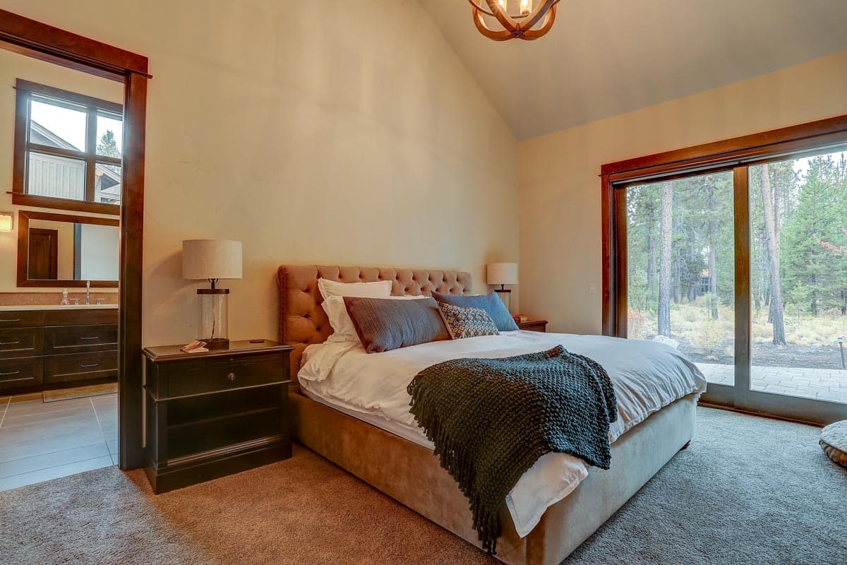 Bedroom with tufted headboard, vaulted ceiling, and sliding glass door overlooking wooded area.