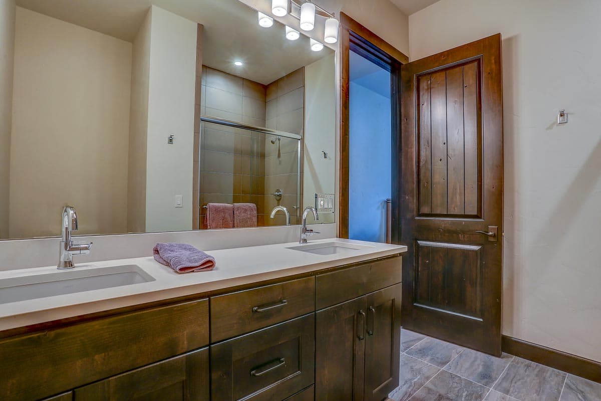 Bathroom vanity with double sinks and dark wood cabinets, reflected shower stall, and rustic wooden door.