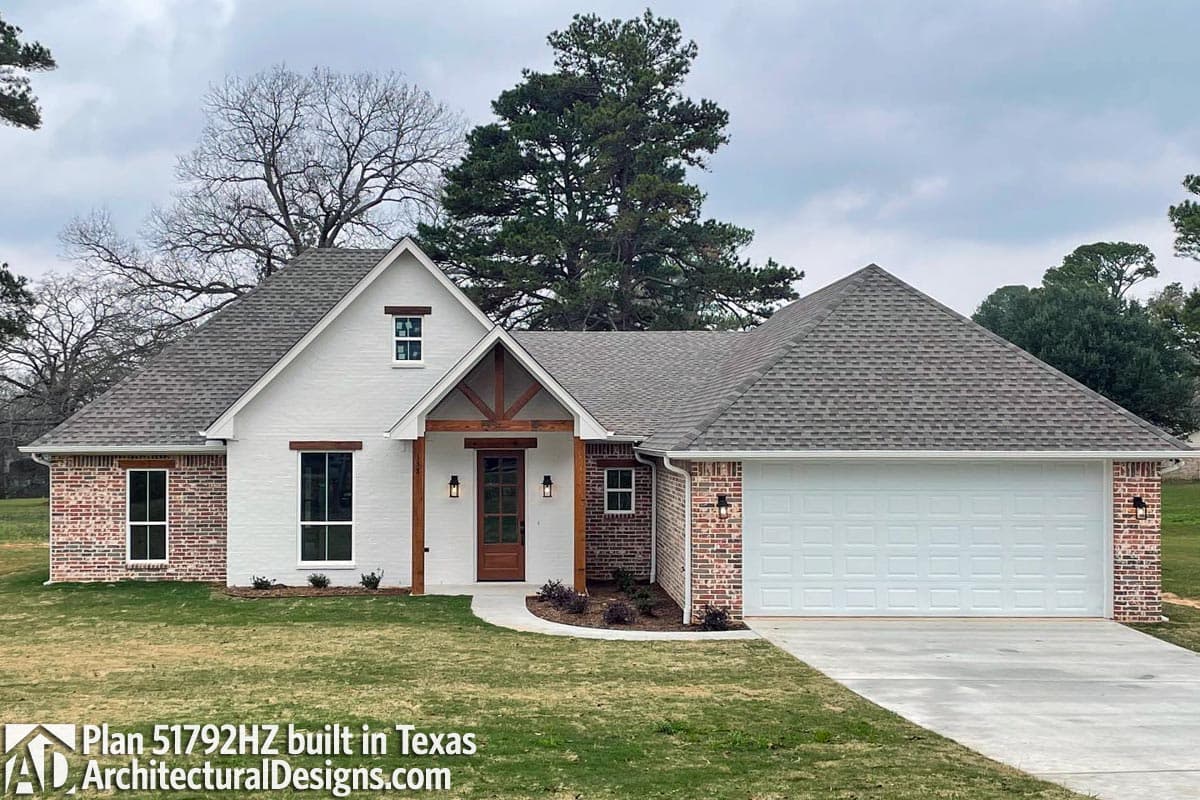 House plan exterior: One-story home with brick accents, prominent gabled entryway with exposed wood beams, and a two-car garage.