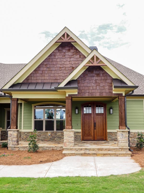 House plan exterior featuring a covered porch with stone bases, wood columns, double wooden doors, and prominent gables with decorative wood bracing.