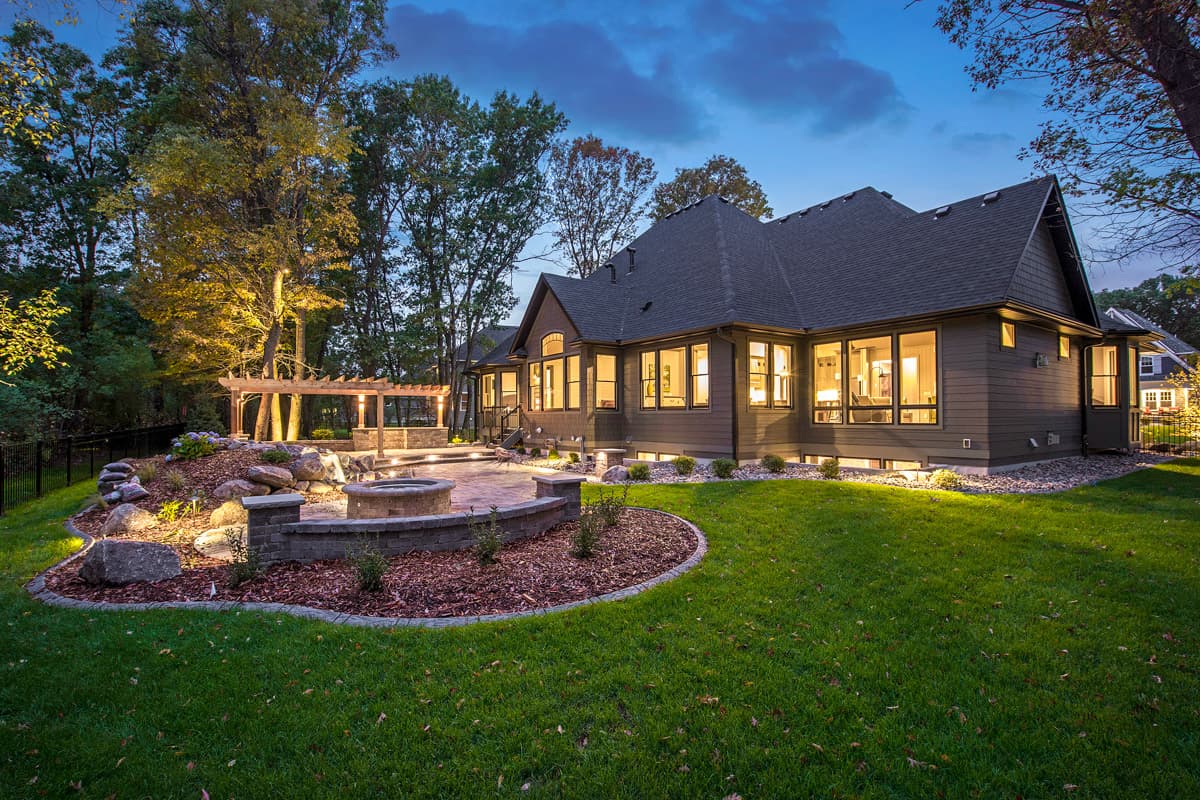 House plan exterior view of a modern home with a dark roof, wide windows, pergola, and stone fire pit patio.