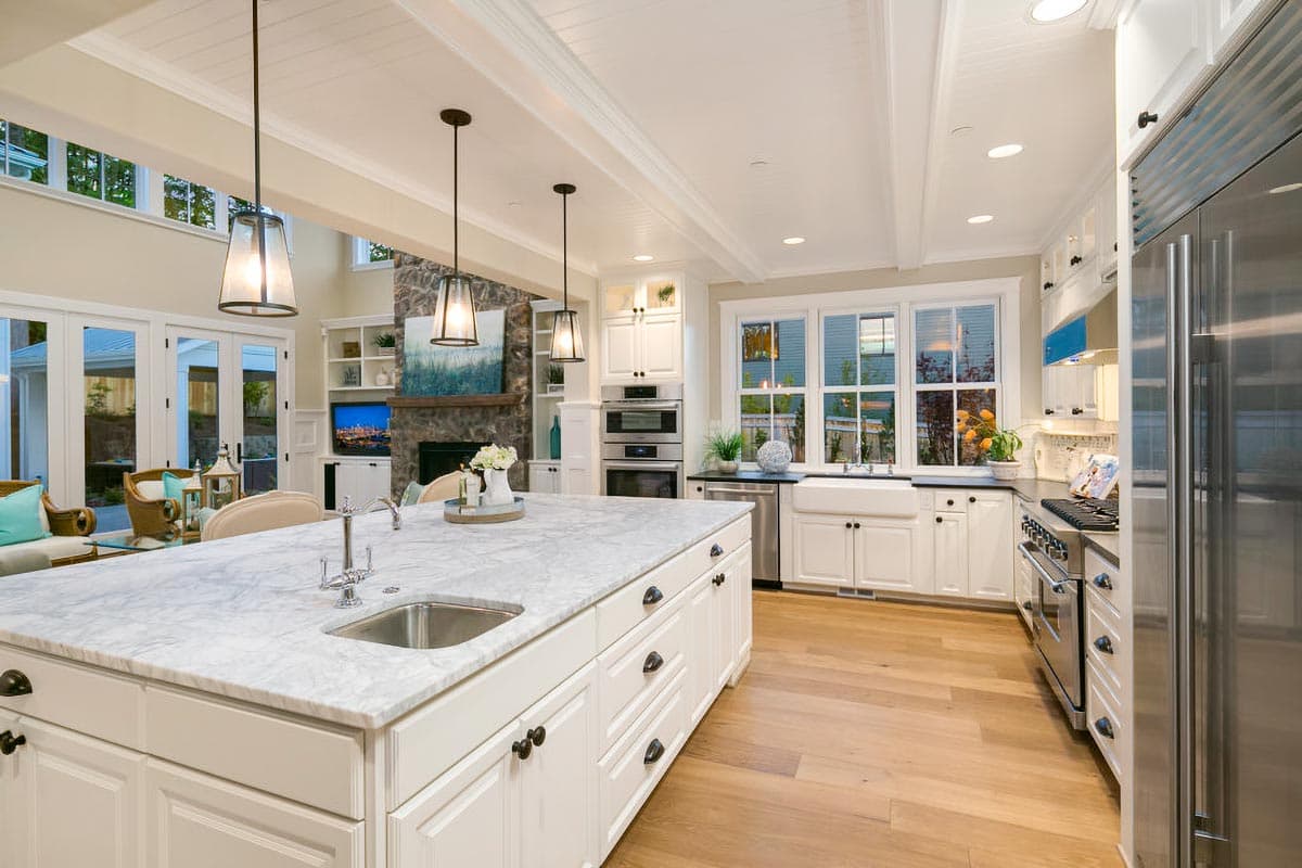 Kitchen with large marble island, white cabinets, farmhouse sink, stainless steel appliances, and coffered ceiling.