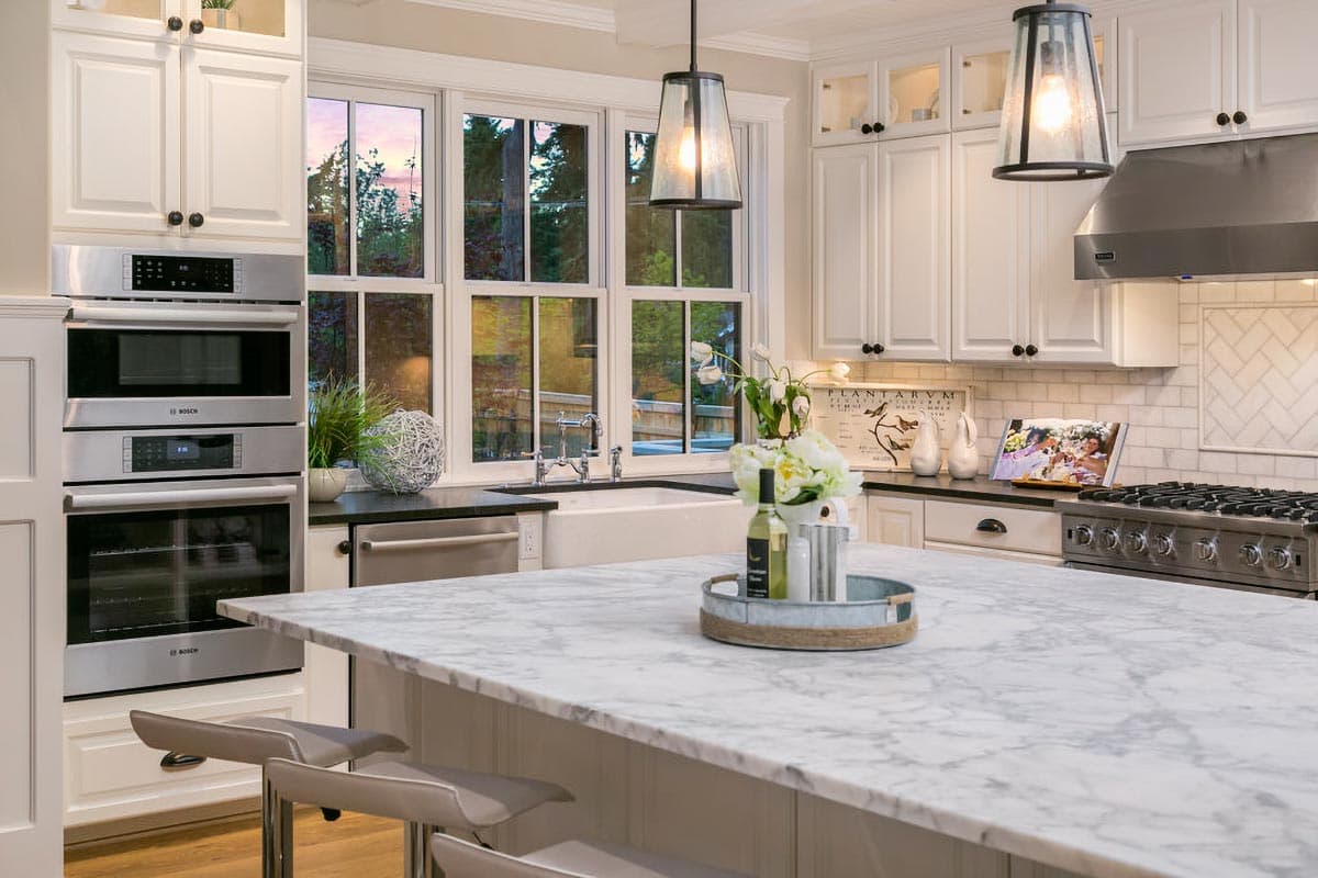 Modern kitchen interior with marble island, white cabinetry, stainless steel appliances, and large windows.