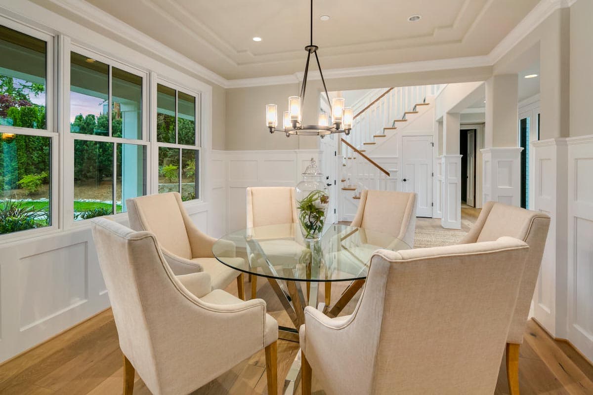 Dining room with glass-top table, upholstered chairs, coffered ceiling, wainscoting, and staircase.