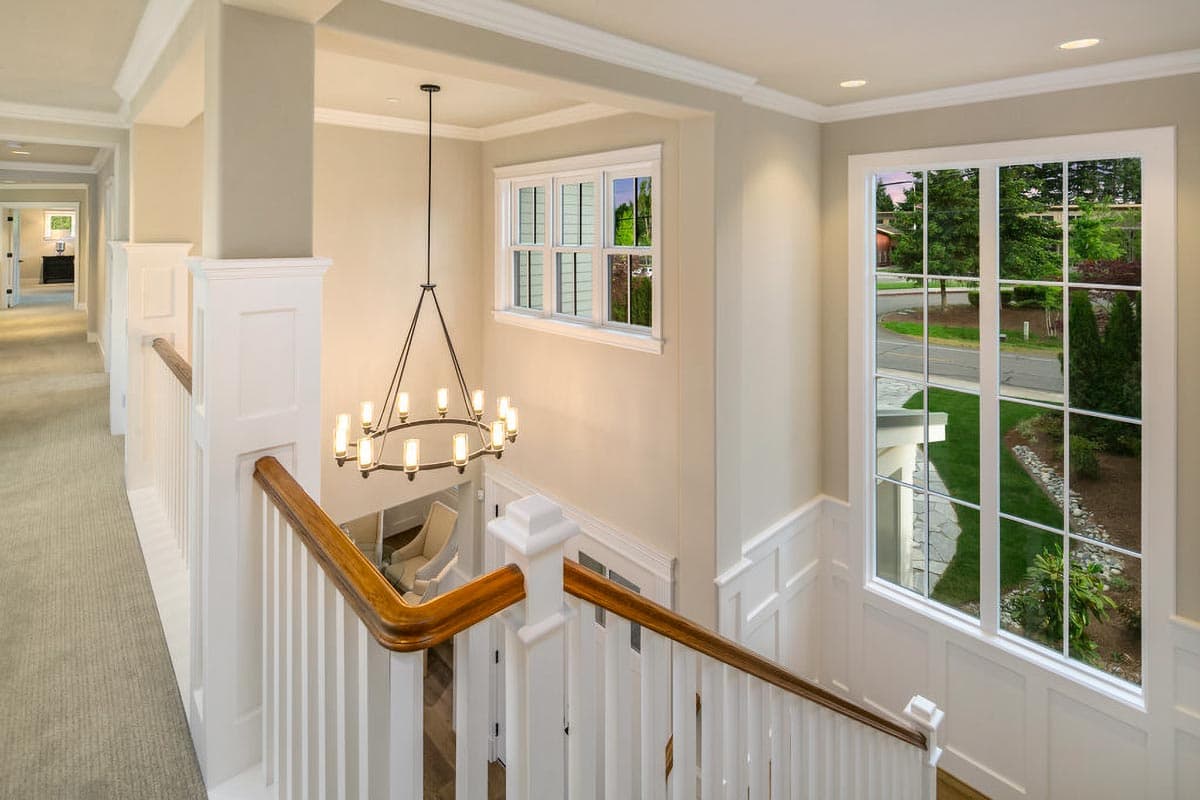 Interior view of a hallway landing with a staircase, chandelier, and large windows overlooking a residential street.