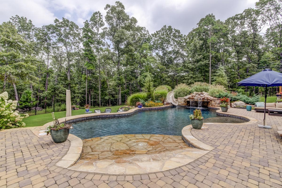 Backyard landscape with curved pool, waterfall, slide, and paved patio seating area surrounded by trees.