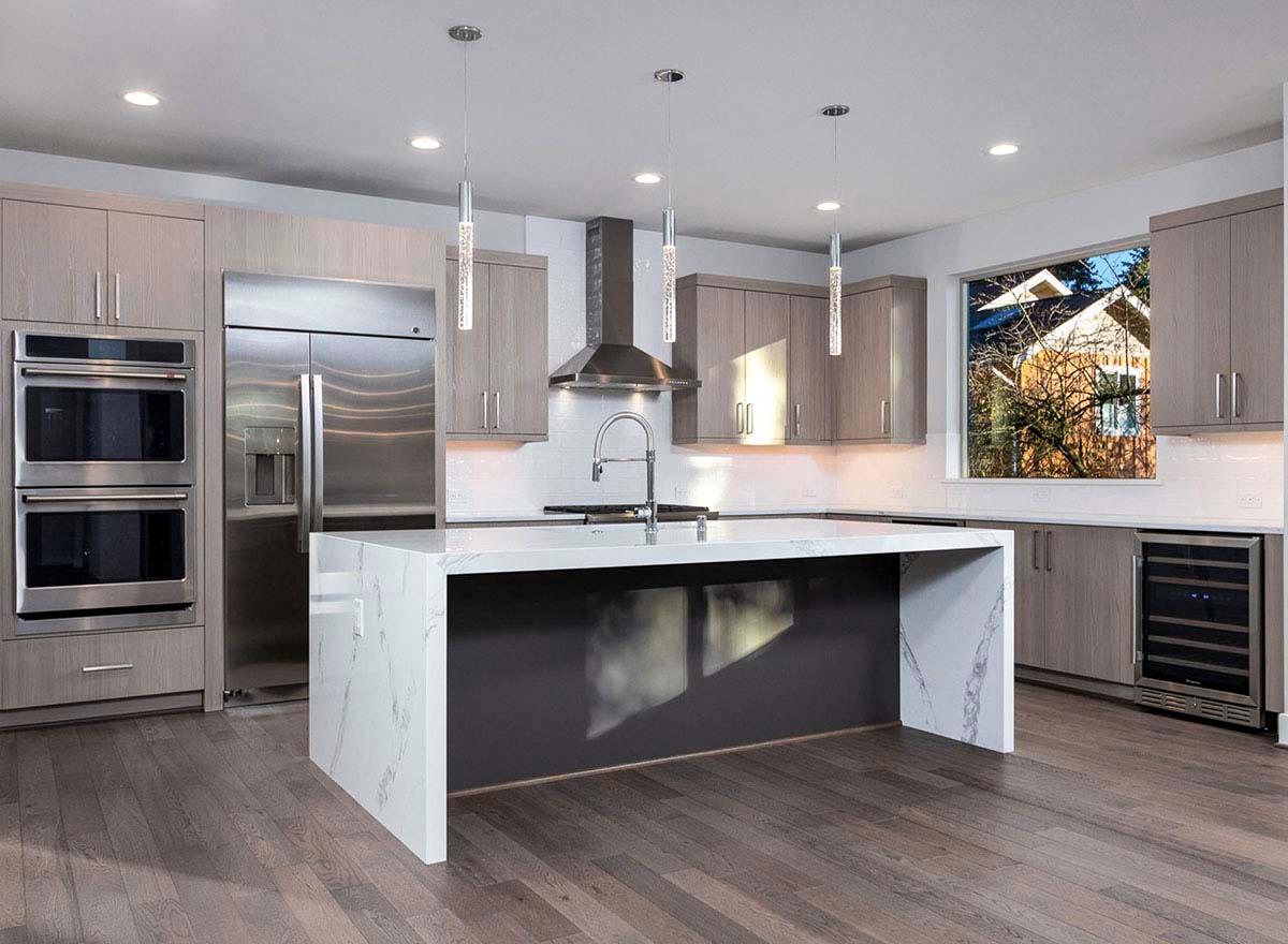Modern kitchen interior with large island, stainless steel appliances, gray wood cabinetry, and a sink with faucet.