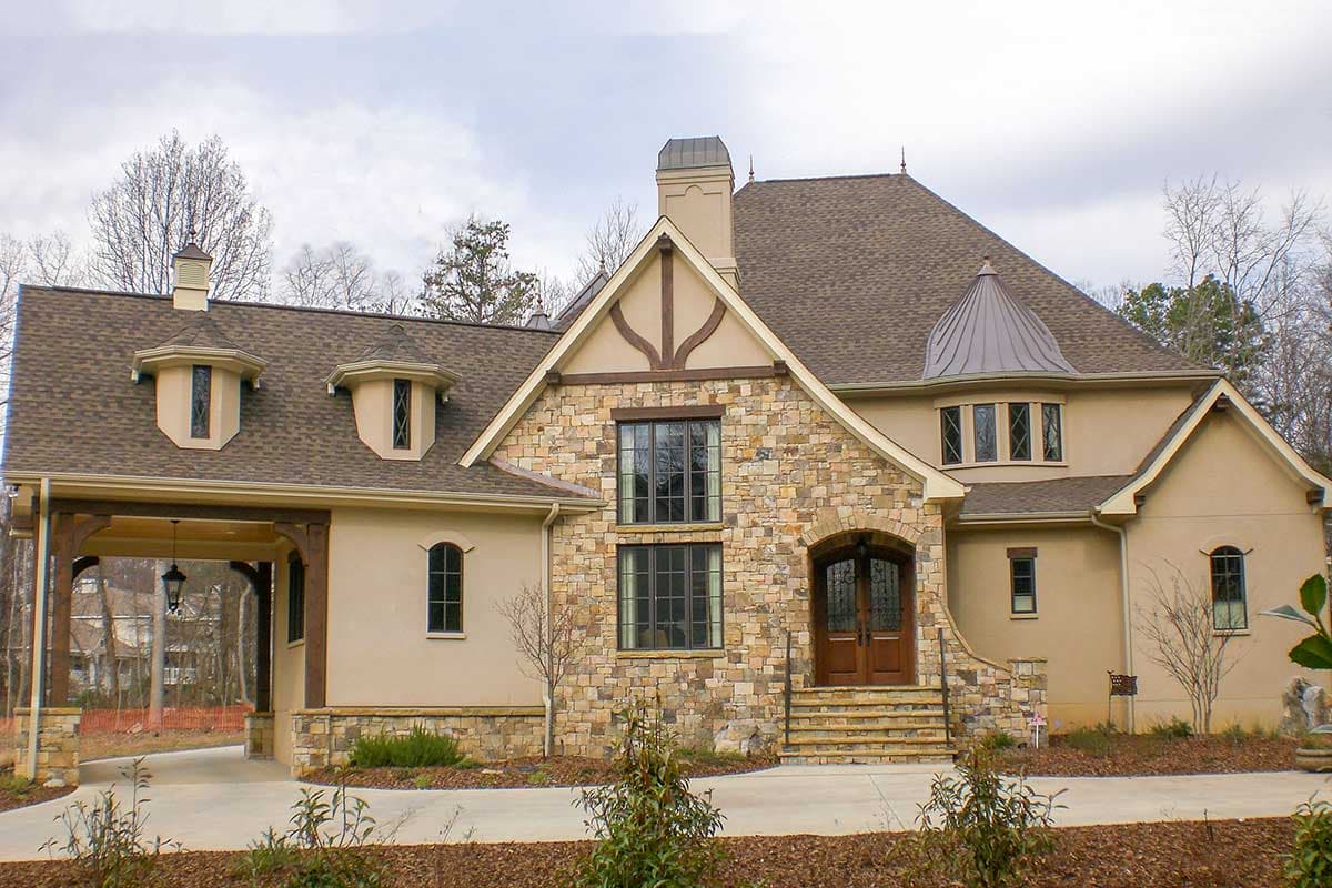 House plan exterior featuring stone facade, multiple gables, and a porte-cochère. Two-story home with a turret roofline and dormers.