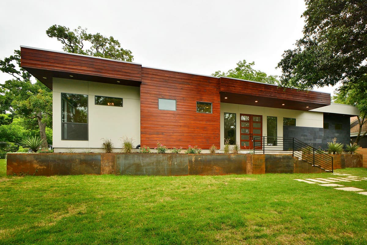 Modern house exterior with wood siding, large windows, flat roof, and elevated entrance with stone accents.