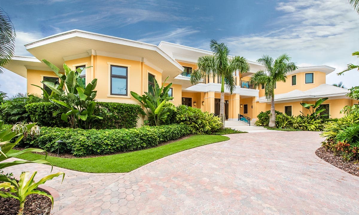 House plan exterior with multi-story structure, flat rooflines, and a covered entryway. Lush landscaping and a circular driveway are visible.