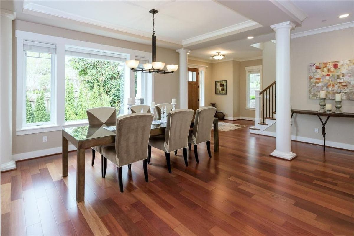 Dining room with a table and chairs, large windows, columns, and staircase, featuring hardwood floors.