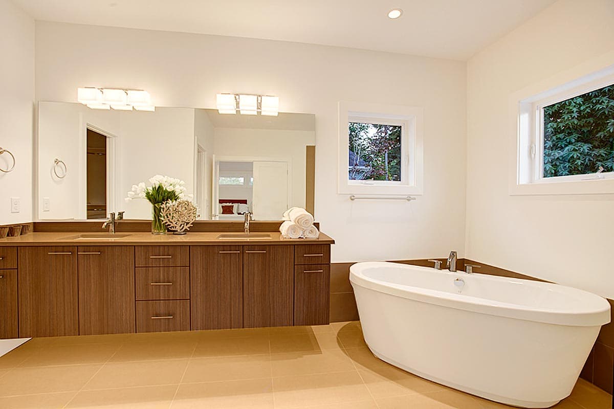 Modern bathroom with floating wood vanity, dual sinks, freestanding tub, and two windows with green foliage views.