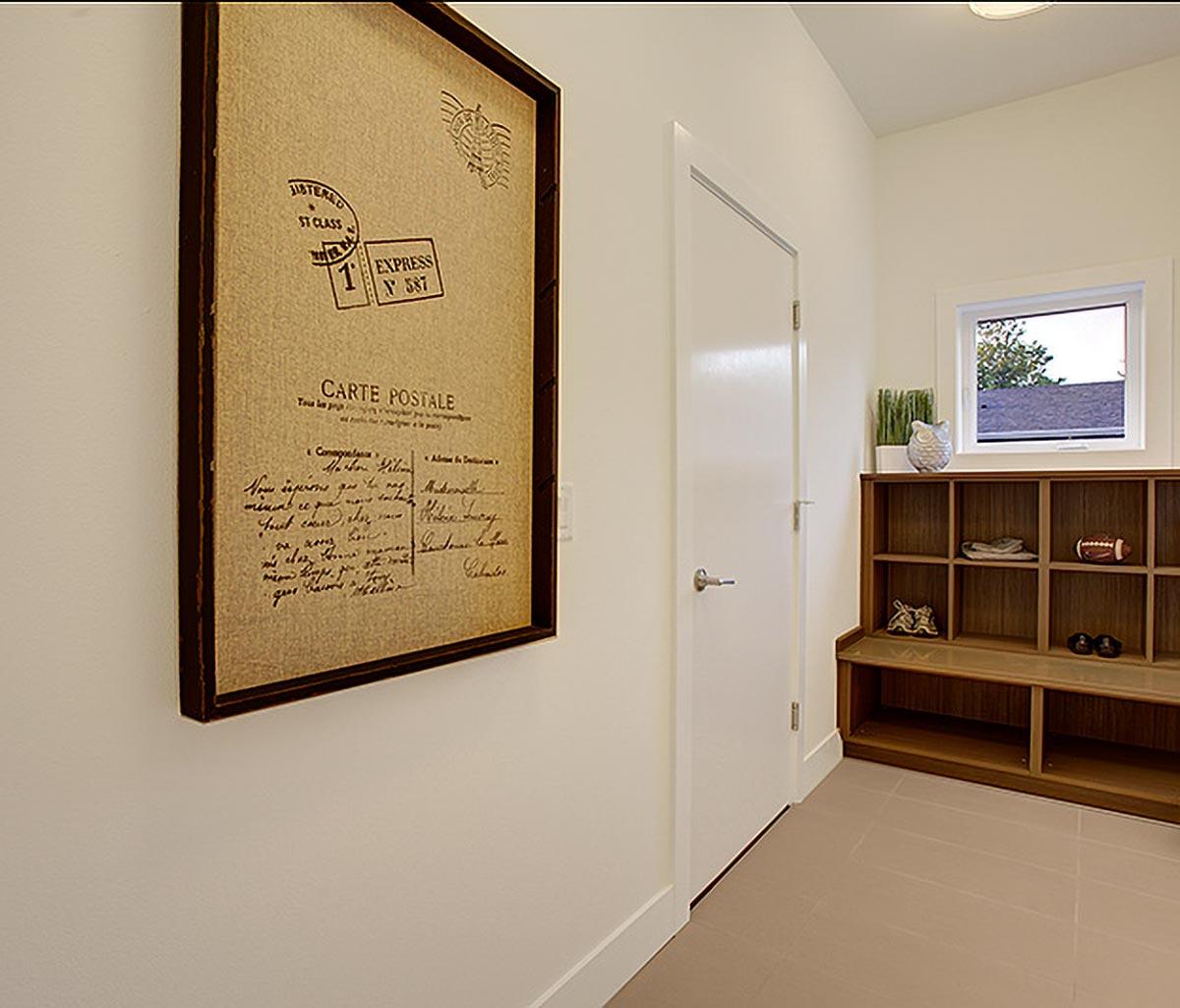 Interior view of a mudroom with a framed postcard, a white door, and a wooden cubby bench with a window above.