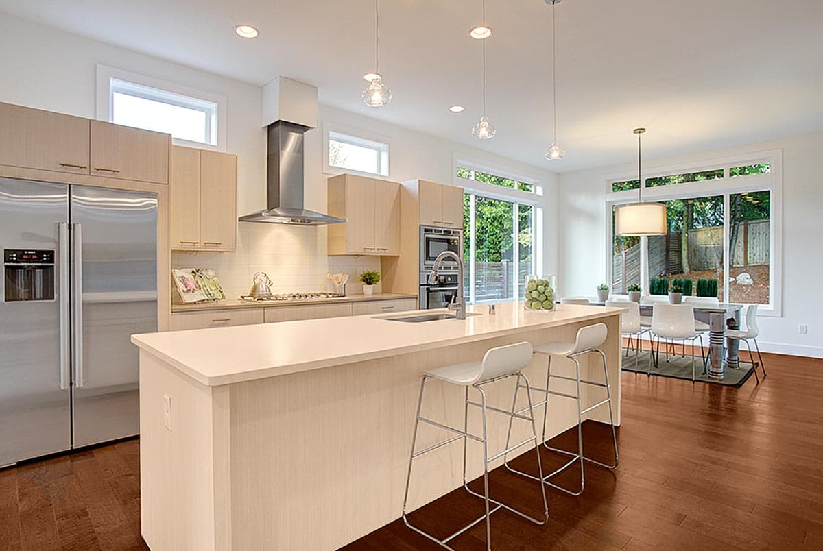 Modern kitchen featuring a large island with seating, stainless steel appliances, light wood cabinetry, and dining area with large windows.