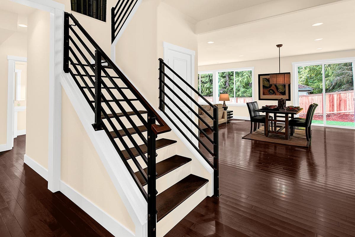 Interior view of a house showing a staircase with black metal railings, open-concept living and dining areas with dark wood floors and large windows.