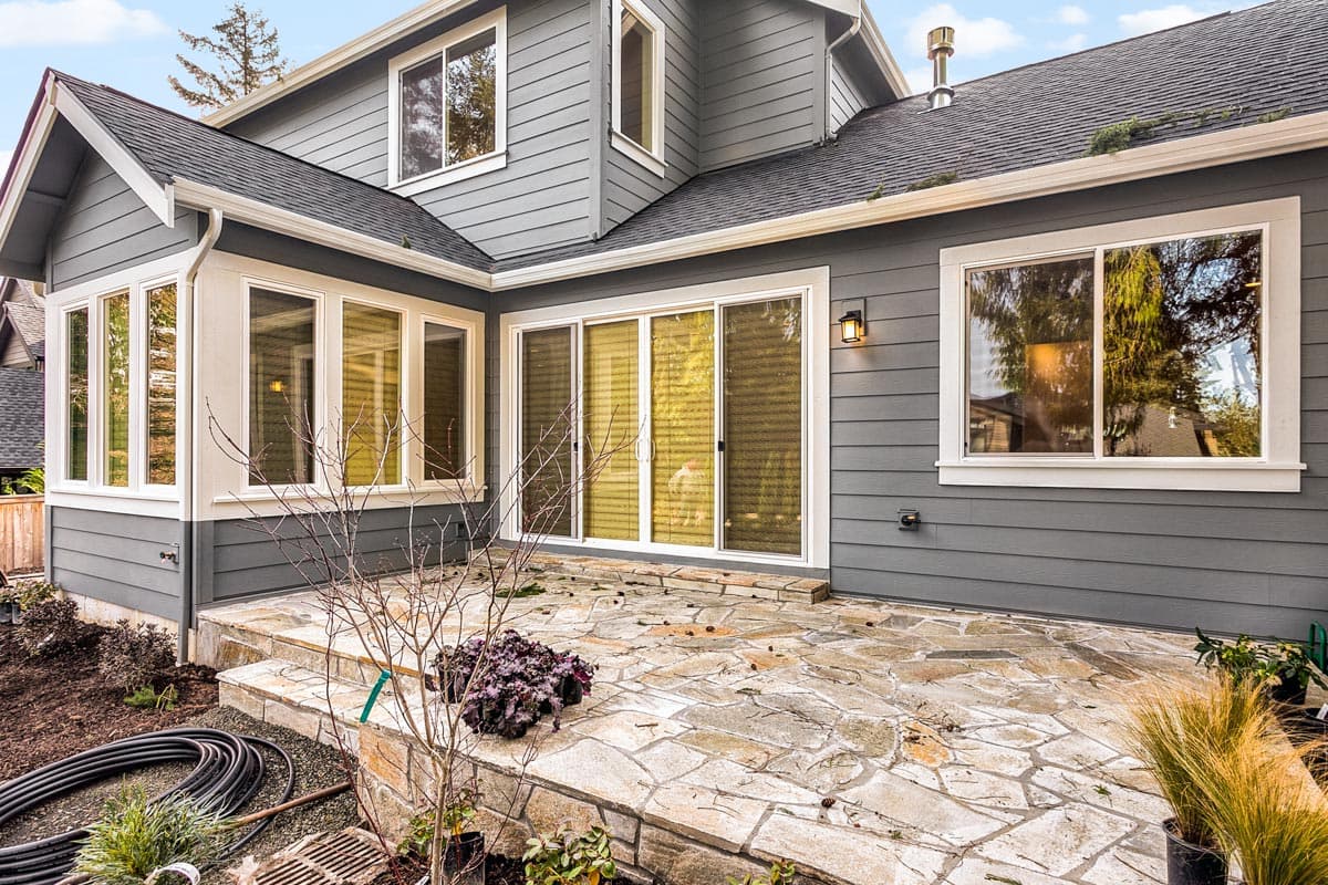 House plan exterior with gray siding, gabled dormer, multiple windows, sliding glass doors, and a flagstone patio.