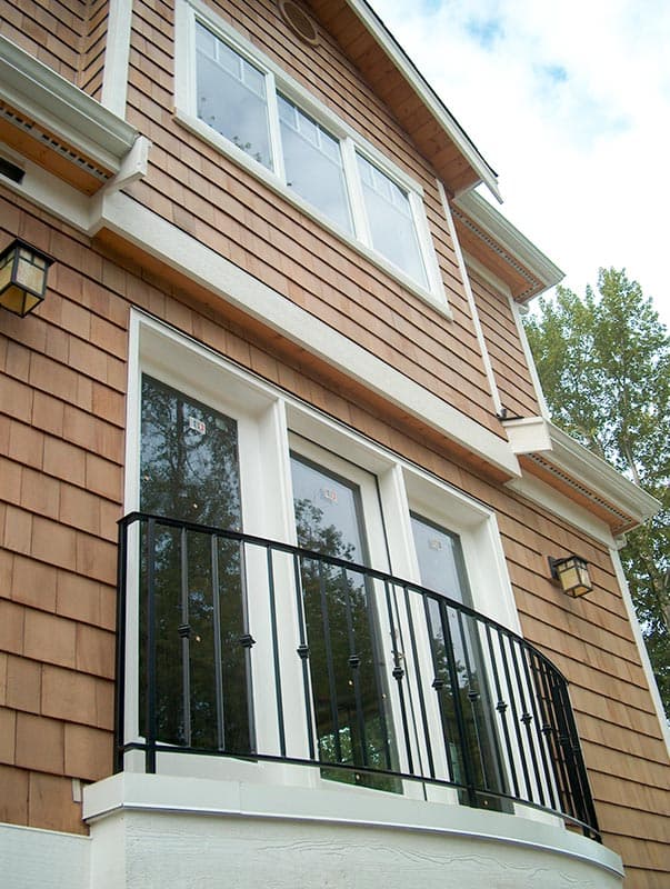 House plan exterior detail with cedar shake siding, white trim, multi-pane windows, French doors, and a curved black metal balcony railing.