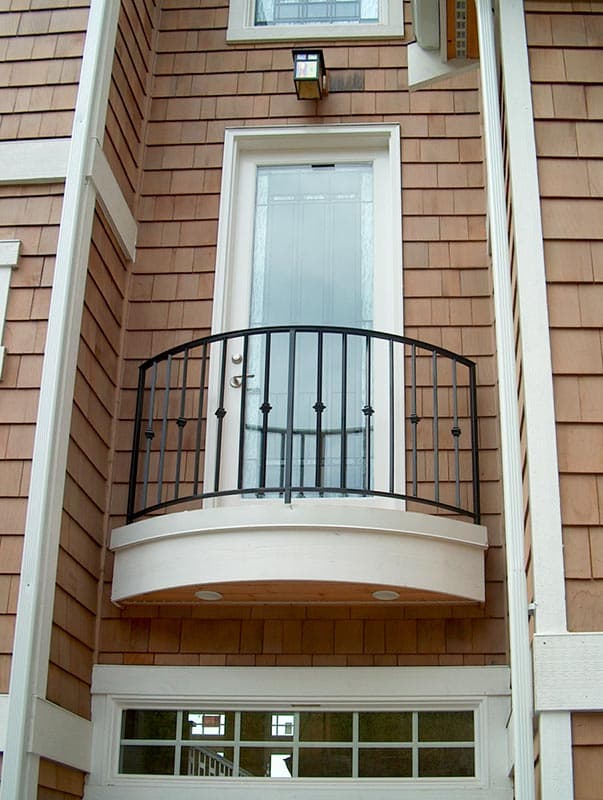 Exterior detail showing shingle siding, balcony with wrought iron railing, and a glass-paneled door above a multi-pane window.