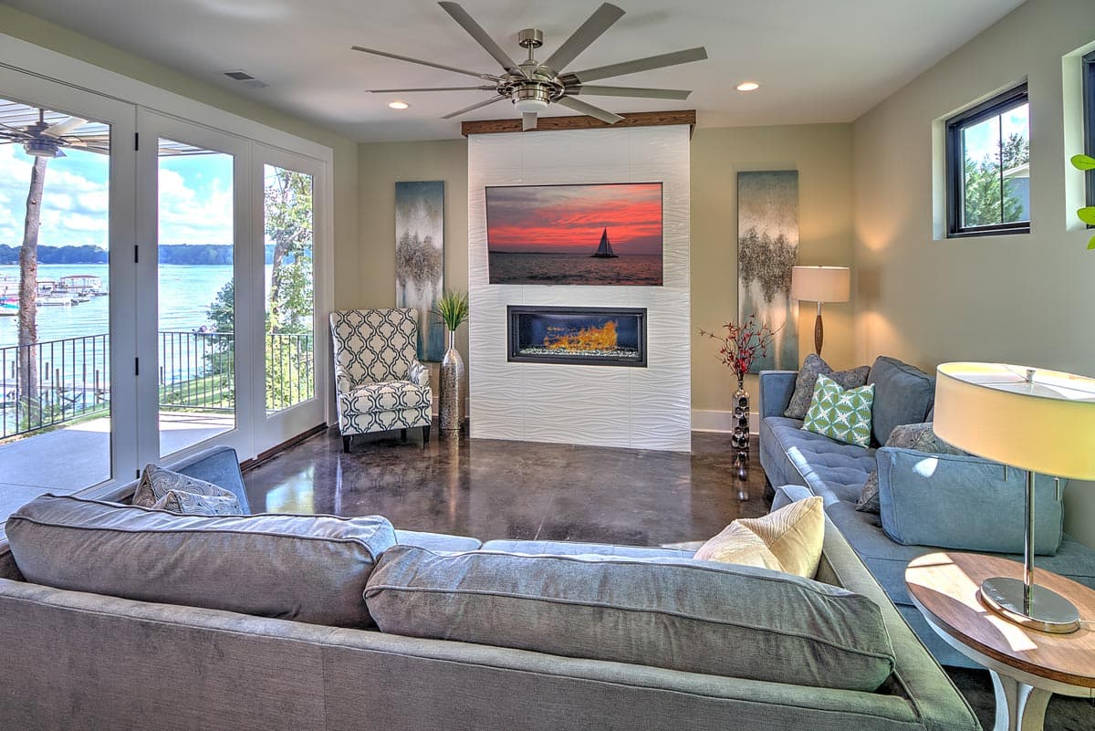 Living room with fireplace, TV, large windows, and water view. Features modern furniture and a ceiling fan.