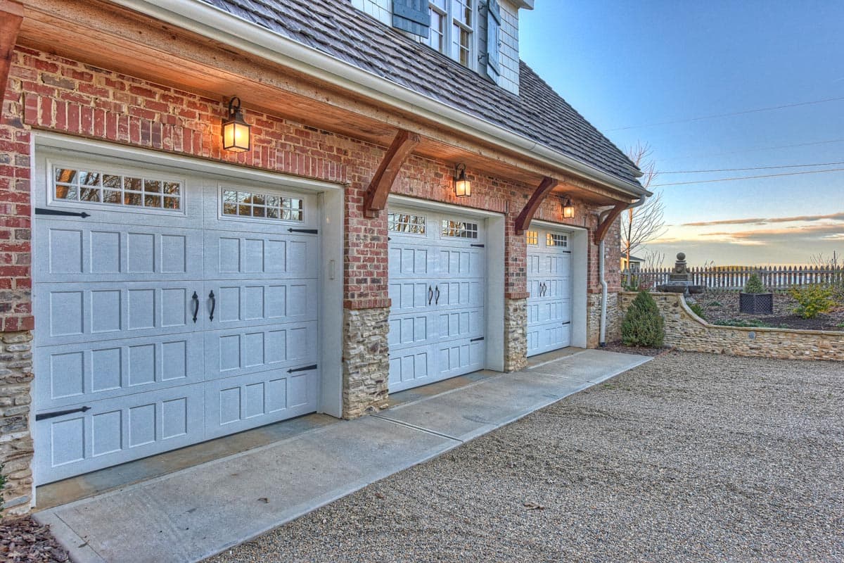 Three-car garage exterior with brick facade, stone accents, wooden beams, and glass-paneled doors.