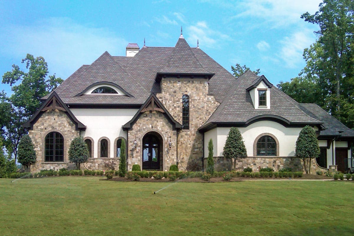 House plan exterior with stone and stucco facade, multi-gabled roof, arched entry, and dormers. Features include a large oval window.