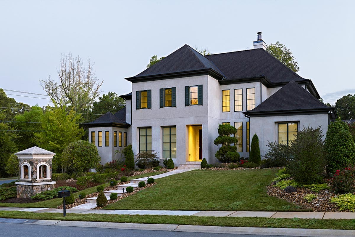 Two-story house plan exterior with stucco finish, dark pitched roof, multiple gables, and varied window styles.