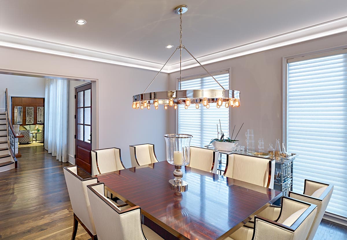 Interior view of a dining room with a polished wood table, upholstered chairs, a circular chandelier, and large windows.